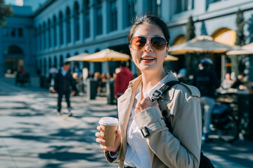 Stylish happy young asian japanese woman wearing sunglasses face camera smiling. girl holding coffee to go while standing on street. charming elegant lady carry bag walking by sunny outdoor cafe shop