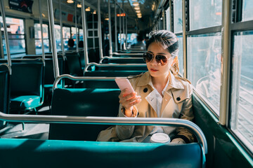 elegant businesswoman in sunglasses sending text message on mobile phone in bus. beautiful female commuter holding cup of coffee on way to work in morning. office lady using cellphone online.