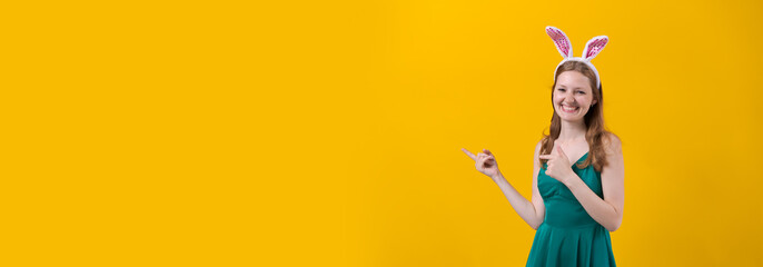 Positive cute girl posing with bunny ears and in green dress on yellow background in the studio. Caucasian young woman joyfully meets easter. Spring mood gestures with hands in different directions