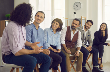 Wall Mural - Group of happy interested business people listening to a coworker sitting in the office during a meeting. Creative young man talking to team members and telling a story about professional motivation