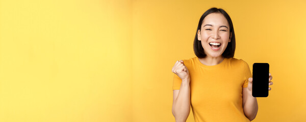 Wall Mural - Excited asian woman, showing smartphone app and triumphing, celebrating on mobile phone, standing over yellow background