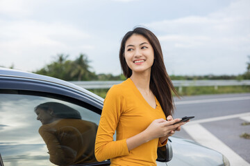 A young Asian woman drives a beautiful nature drive. she was standing in front of the car on the roadside. she uses the smartphone to call services.