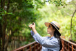 © Chanakon - Asian woman travel using cell phone taking video and photo in nature with sunlight. Traveller blogger in mangrove forest. Rest on vacation holiday weekend. Beautiful female with hat and blue shirt