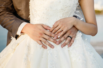 Wall Mural - Groom hugging beautiful bride in a white dress from behind Hands Close up Husband and wife who is pregnant posing by hugging each other