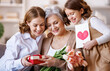 © JenkoAtaman - Happy International Women's Day.Smiling  daughter and granddaughter giving flowers  and gift to grandmother   celebrate spring holiday Mother's Day at home