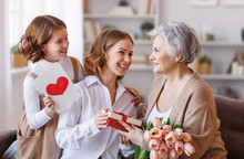 Happy International Women's Day.Smiling  Daughter And Granddaughter Giving Flowers  And Gift To Grandmother   Celebrate Spring Holiday Mother's Day At Home