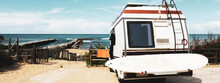 Horizontal Banner Or Header With Rear View Of Vintage Camper Parked On The Beach Against A Scenic View - Caravan Of Surfer With A Surfboard On Back - Nomadic And Van Life Concept