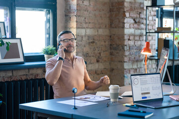 Wall Mural - Businessman in T-shirt talking on mobile phone while sitting at the table with computer and waiting for his colleagues at meeting