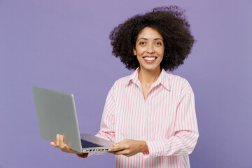 Wall Mural - Young smiling happy satisfied fun woman of African American ethnicity 20s wear pink striped shirt hold use work on laptop pc computer isolated on plain pastel light purple background studio portrait.