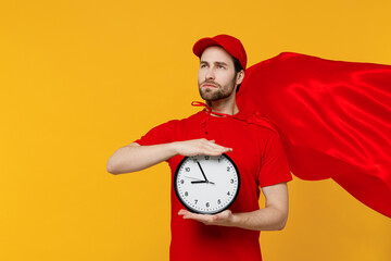 Professional minded delivery guy employee man 20s in red cap T-shirt uniform red cloak workwear work as dealer courier hold clock isolated on plain yellow background studio portrait. Service concept.