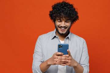 Smiling exultant joyful young bearded Indian man 20s years old wearsblue shirt hold in hand use mobile cell phone typing browsing chatting send sms isolated on plain orange background studio portrait