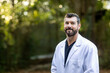A doctor with dark hair and a beard in a white lab coat standing outside in a natural green environment