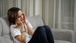 © lenblr - Brunette woman in white t-shirt handles strong headache after hard working day sitting on sofa in living room at home in late evening.