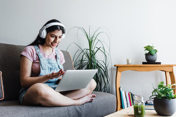 young latin Woman student with headphones using computer in a video call or online class while sitting on sofa at home in Mexico	