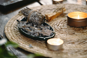 Woman hands burning white sage, before ritual on the table with candles and green plants. Smoke of smudging treats pain and stress, clear negative energy and meditation