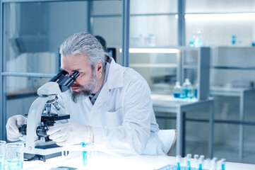 Mature bearded scientist examining the analysis in test tube with microscope sitting at the table in lab