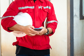 Wall Mural - An engineer or manager in red uniform is holding smartphone and white safety helmet during take group meeting with team staff. Industrial working people in action photo. Selective focus at hand part.