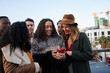 © StratfordProductions - Diverse group of young adult friends on a rooftop terrace in the city, looking and laughing at a photo of biracial girl's cellphone.
