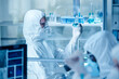 © AnnaStills - Laboratory technician in protective suit examining the blue liquid in flask while working with colleagues in laboratory in danger zone