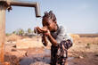 Young black girl greedily drinking fresh water from a pipe somewhere in an African dryland; global warming concept