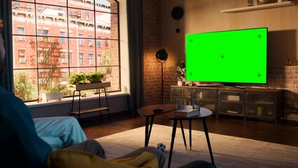 Wall Mural - Young Couple Spending Time at Home, Sitting on a Couch and Watching TV with Green Screen Mock Up Display in Their Stylish Loft Apartment. Man and Woman Streaming Movie or Show and Eating Popcorn.