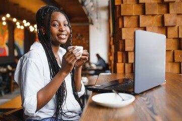 Head shot portrait of happy smiling African American woman sitting at table in cafe, looking at camera, excited female posing, working at computer, doing homework, preparing report in coffee house