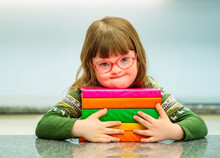 A Child With Down Syndrome With Glasses Sitting At A Table With Books In An Embrace