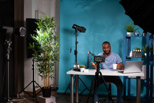 Content Creator Talking To Followers In In Front Of Filming Video Camera On Tripod Sitting At Desk Holding Cup. Wide View Of Vlogging Studio Setup With Smiling Vlogger Interacting With Audience.