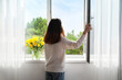 © Pixel-Shot - Pretty young Asian woman opening window at home on sunny day, back view