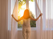 © Pixel-Shot - Pretty young woman opening curtains in room on sunny day, back view