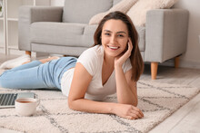 Young Woman With Cup Of Tea And Laptop Lying On Carpet At Home