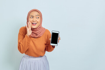 portrait of cheerful young asian muslim woman in brown sweater and headscarf showing blank mobile ph