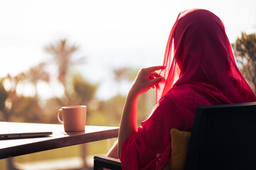 Wall Mural - Silhouette of muslim, arab woman in red head scarf, hijab sitting at the table on terrace with laptop and cup of coffee opposite sunrise palm sea beach