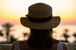 © flowertiare - Silhouette of woman in straw hat standing on balcony and looking at sunset palm sea beach. Rear view