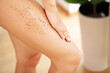 Beautiful young woman applying body scrub on leg at home