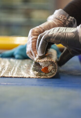 Wall Mural - Shallow focus of hands with gloves rolling a sandwich with cheese and tomatoes on a blue surface