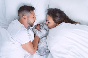 Wall Mural - Young couple lying in the bed with adorable cat. Woman kissing cute cat. Love for cats or pets. Sleep with pets