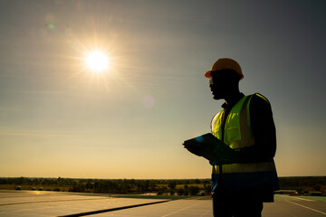 African man engineer using digital tablet maintaining solar cell panels on building rooftop. Technician working outdoor on ecological solar farm construction. Renewable clean energy technology concept