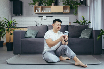 Male asian athlete at home using phone sitting on the floor during morning exercise, athlete in living room