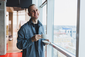 A young man of European appearance in a casual suit drinks coffee and looks out the window