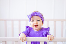 Cute Portrait Of A Baby Girl In A Crib Holding On To The Side In Lilac Clothes And With A Bow On Her Head And Laughing, The Concept Of Children's Goods