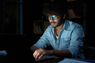 Wall Mural - Focused young indian ethnic business man developer or web designer wearing glasses typing on laptop distance working online late at night at home or in dark office using laptop sitting at desk.