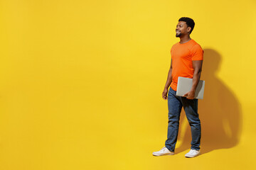 Wall Mural - Full body student fun young man of African American ethnicity 20s wear orange t-shirt hold closed laptop pc computer walk isolated on plain yellow background studio portrait. People lifestyle concept.