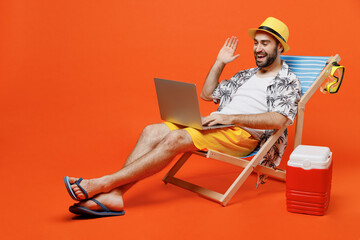 Young tourist man wear beach shirt hat lie on deckchair hold use work on laptop pc computer say hallo hi isolated on plain orange background studio portrait. Summer vacation sea rest sun tan concept