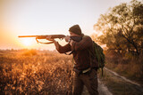 Fototapeta Sport - Young strong soldier with riffle and ammunition belt moving outdoors by the dirt road in sunset time