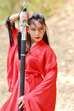 Young beautiful asian woman dressing in traditional Chinese old fashion warrior style with ancient word. Cute girl in red dress looking away in nature outdoor. Travel in Asia concept