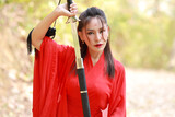 Young beautiful asian woman dressing in traditional Chinese old fashion warrior style with ancient word. Cute girl in red dress looking away in nature outdoor. Travel in Asia concept