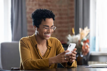 Happy African millennial business woman in eyeglasses using online app on smartphone at home office workplace table, reading text message, tying, chatting on internet, making video call