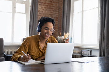 Cheerful Black college student in earphones attending virtual class, writing notes, watching webinar on Internet, studying online. African worker in headphones talking to customer on video call