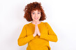 © Jihan - young redhead girl wearing yellow sweater over white background keeps palms together, has pleased expression. Glad attractive male makes request, pleads for mercy. Hopeful young adult.